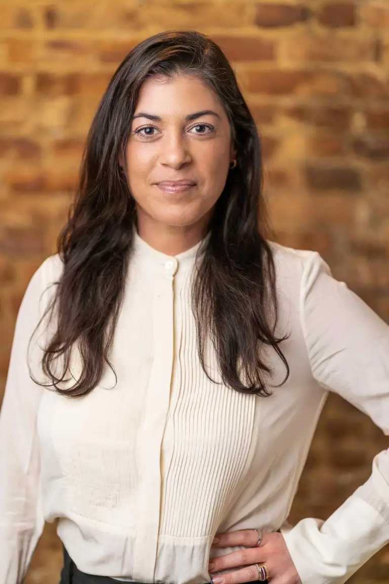 Professional portrait of a woman with long dark hair wearing a white blouse, standing against a brick wall.