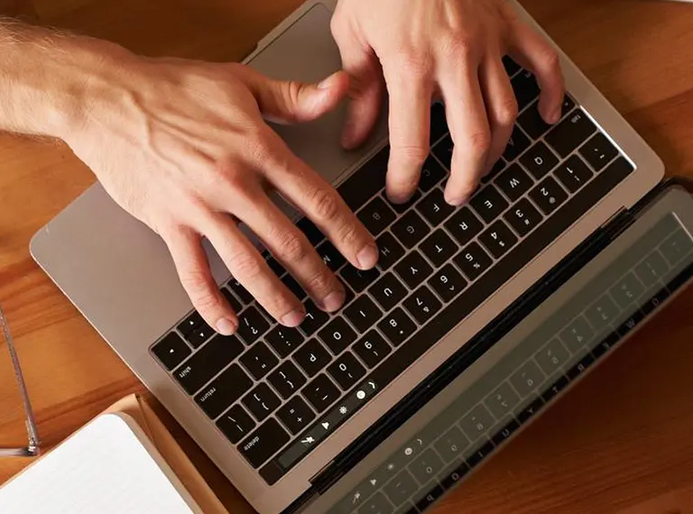 Close-up of hands typing on a silver laptop keyboard on a wooden desk.