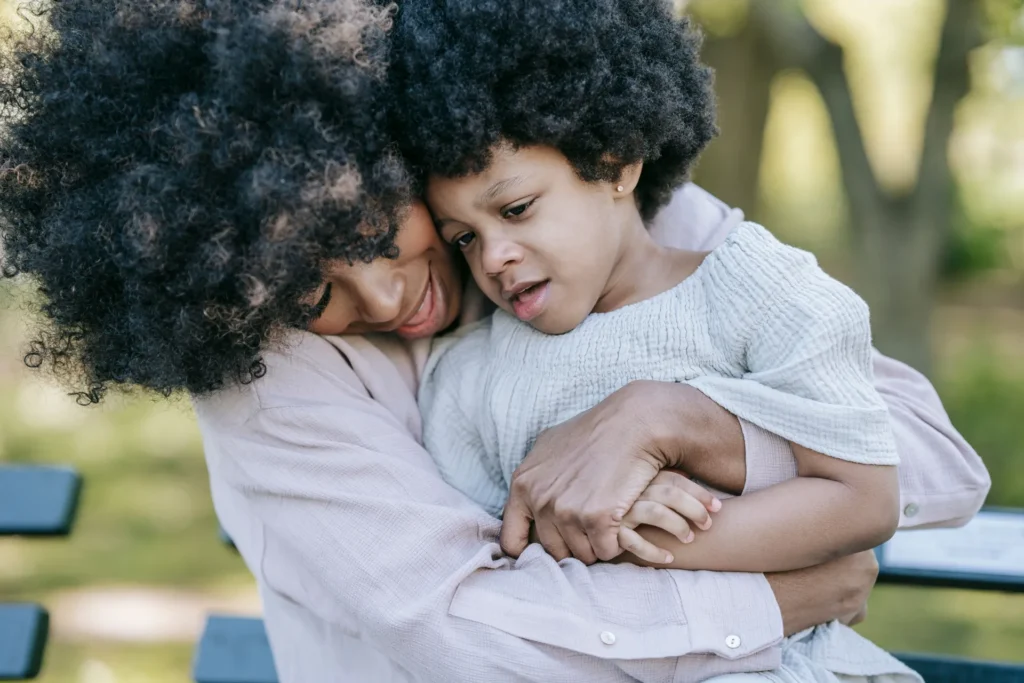 Adult and child with curly hair embracing outdoors, both wearing light-colored clothing.