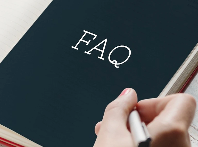 Close-up of an open notebook with "FAQ" text on a dark page and a hand holding a pen.