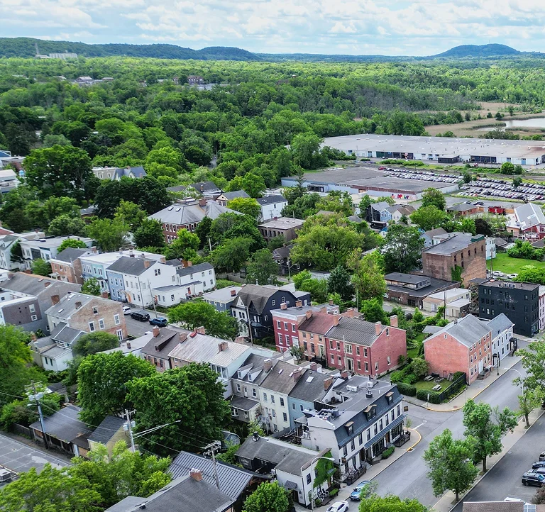 Aerial view of a small town with residential buildings, green trees, and distant hills under a cloudy sky.