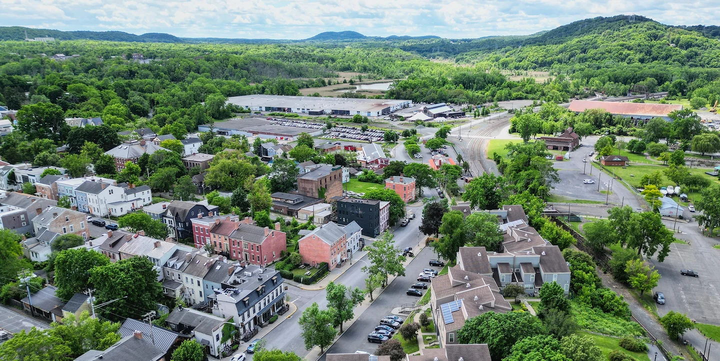 Aerial view of a small town with residential buildings, green trees, parking lots, and distant hills under a partly cloudy sky.