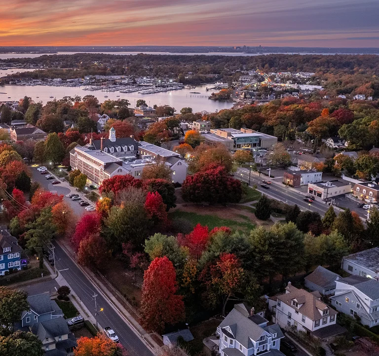 Aerial view of a coastal town at sunset with autumn foliage, buildings, roads, and a marina.