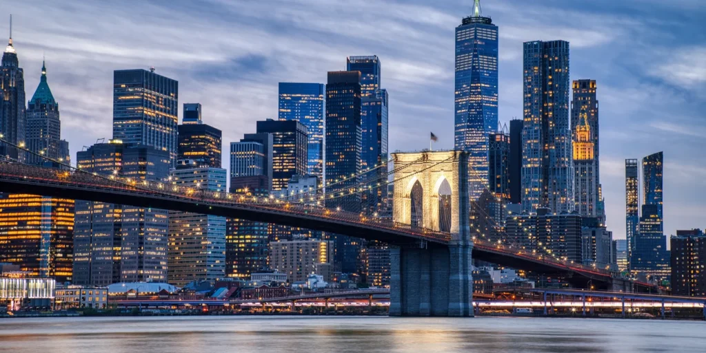 Evening view of Brooklyn Bridge with illuminated cables and New York City skyline in background.
