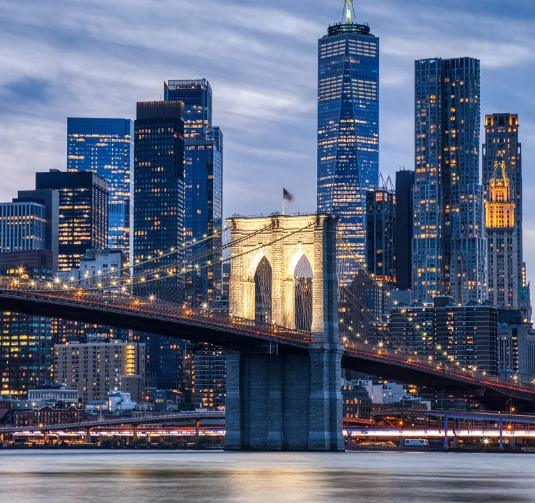 Illuminated Brooklyn Bridge spanning East River with Manhattan skyscrapers at dusk.