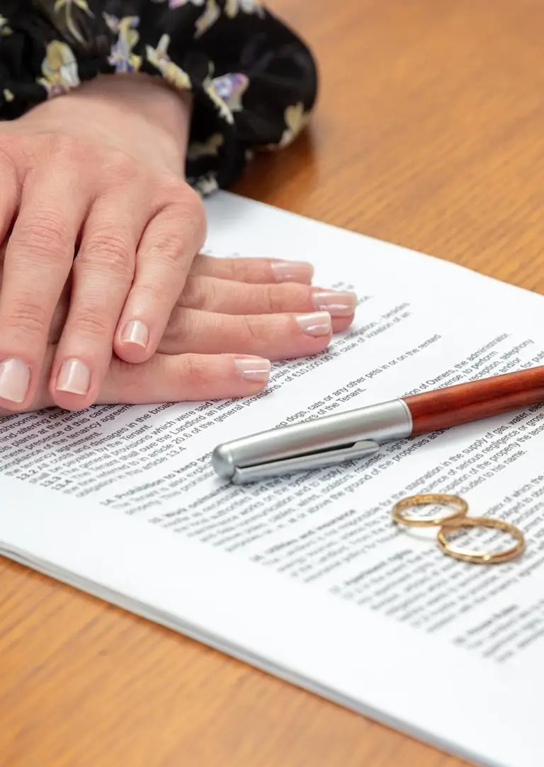 Close-up of hands resting on a legal document with a pen and two gold wedding rings on a wooden table.