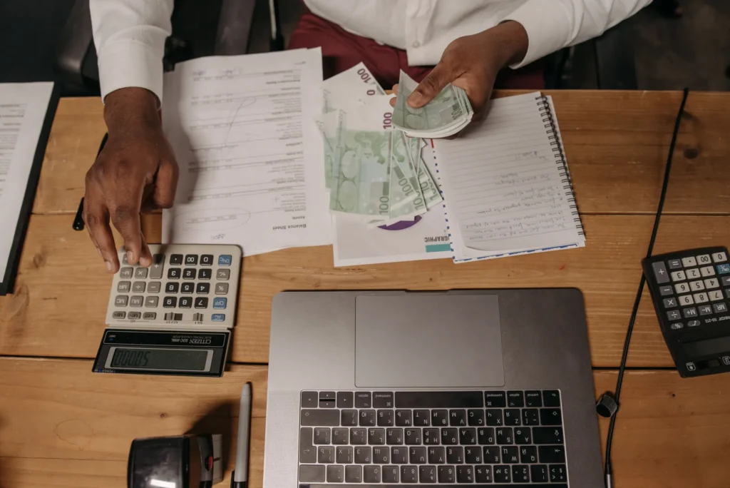Person counting euro banknotes with calculator, documents, and laptop on wooden desk.