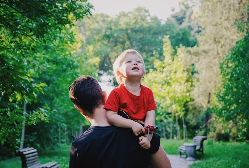 Adult carrying a young child in a red shirt, walking in a green park with benches.
