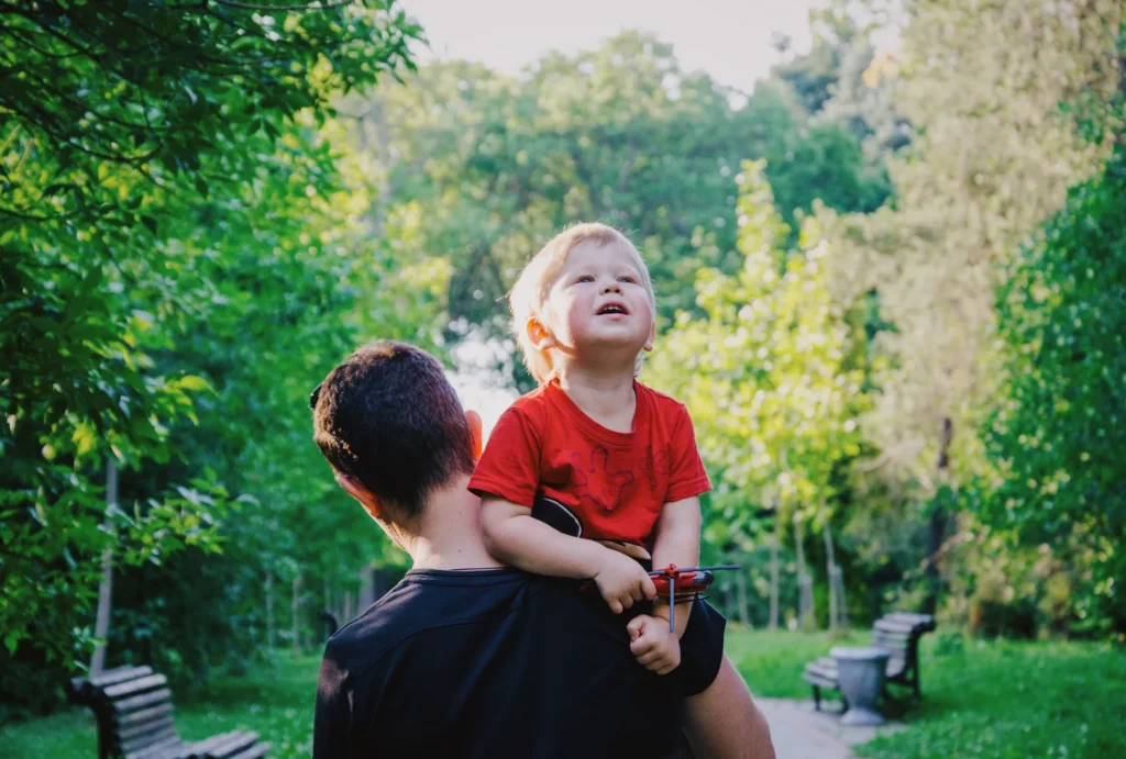 Adult carrying young child in red shirt outdoors, surrounded by green trees and park benches.