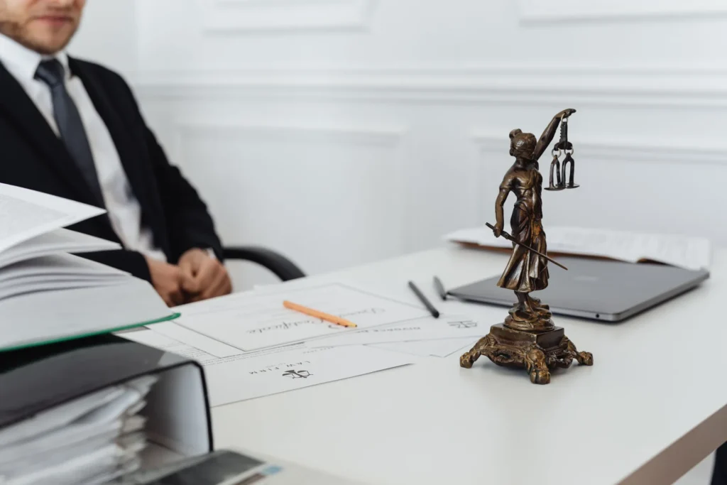 Bronze Lady Justice statue on white desk with legal documents and man in suit in background.