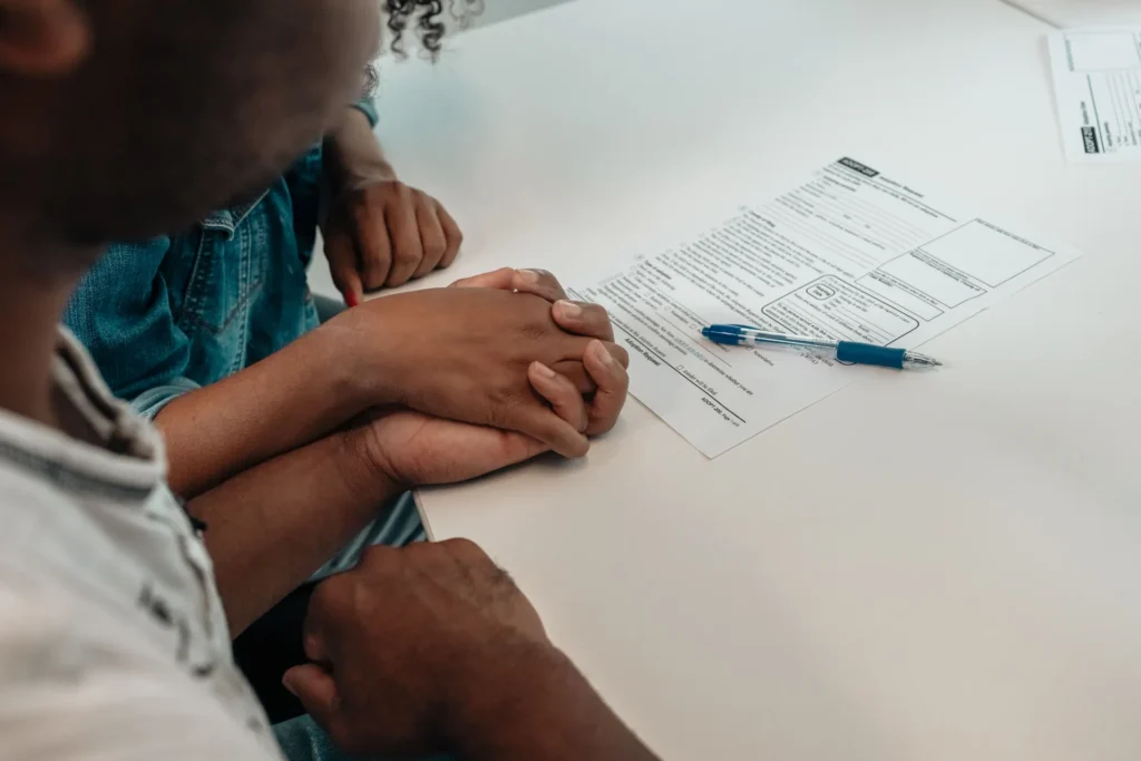 Two individuals holding hands over a white table with a printed document and blue pen.