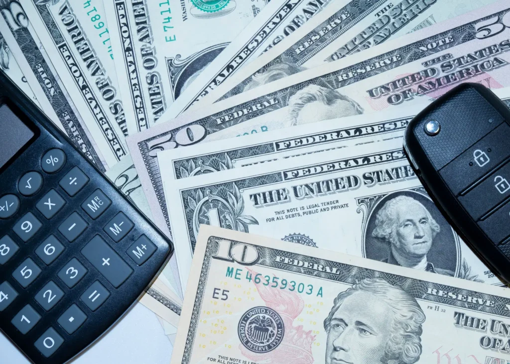 Calculator, US dollar bills of various denominations, and a black car key fob on a white surface.