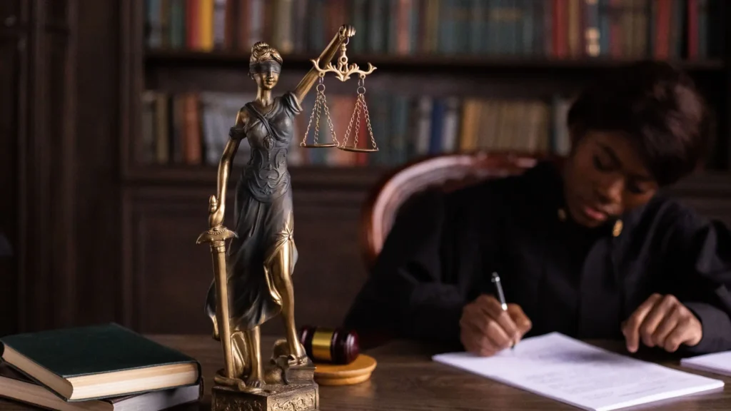 Bronze Lady Justice statue with scales and sword on desk, woman writing in background.