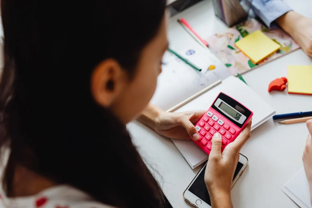 Person holding a pink calculator displaying number 56, with notebooks and stationery on white table.