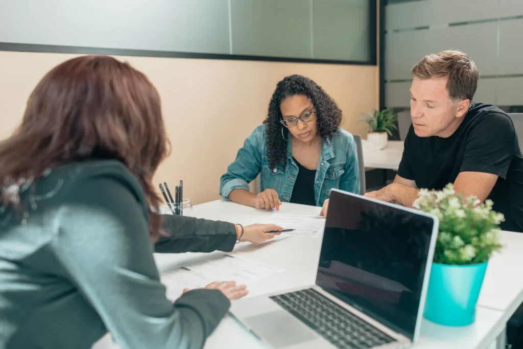 Three professionals reviewing documents at a white table with a laptop and potted plant.