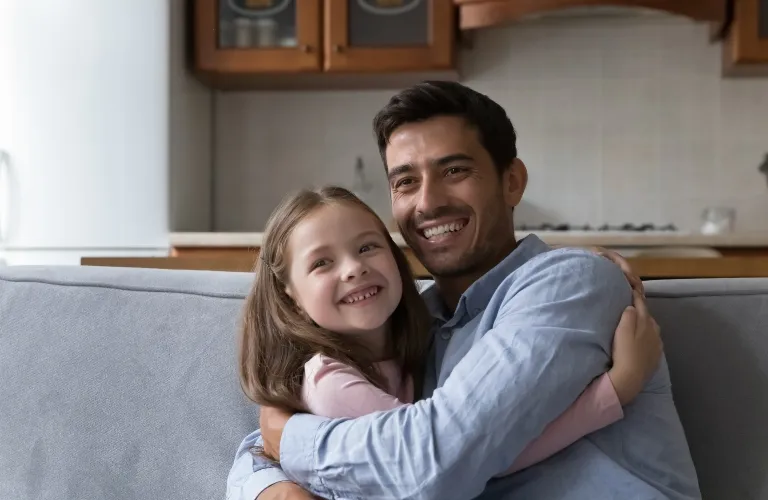 Man and young girl smiling and embracing while seated on a gray couch in a kitchen.
