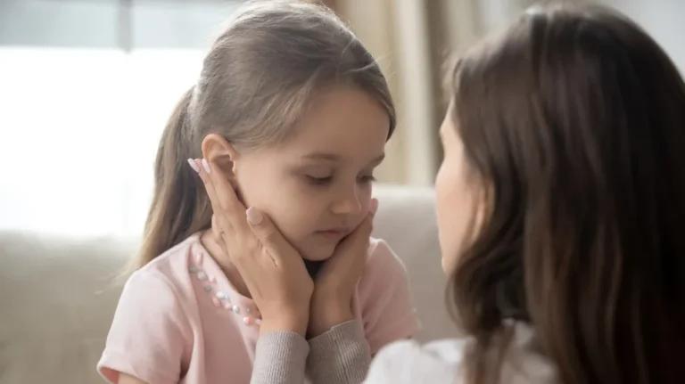 Woman comforting young girl with hands on her cheeks in a softly lit room.