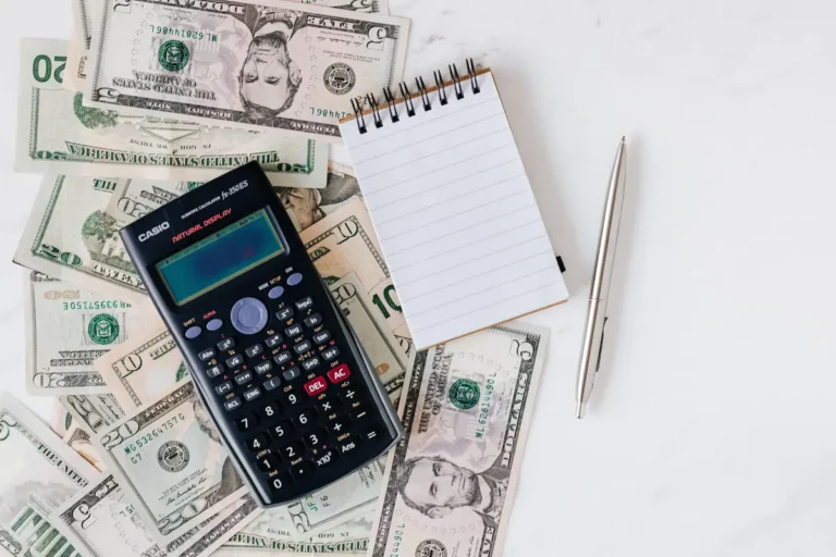 Scientific calculator, blank spiral notepad, silver pen, and scattered U.S. dollar bills on white surface.