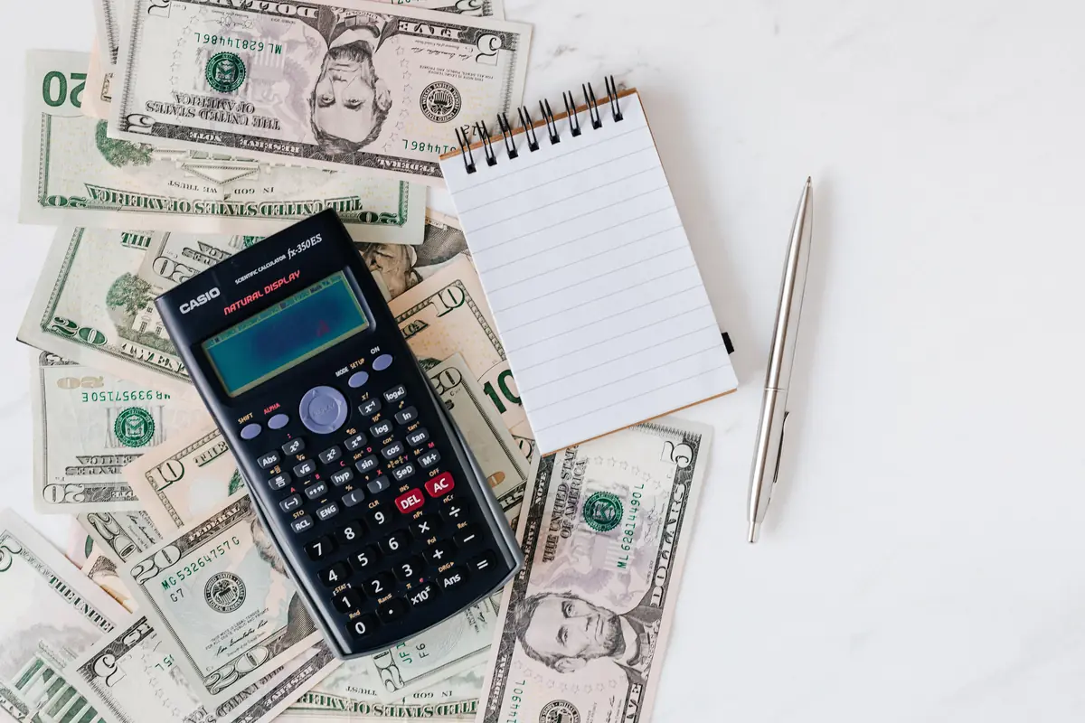 Scientific calculator, blank spiral notepad, silver pen, and scattered U.S. dollar bills on white surface.