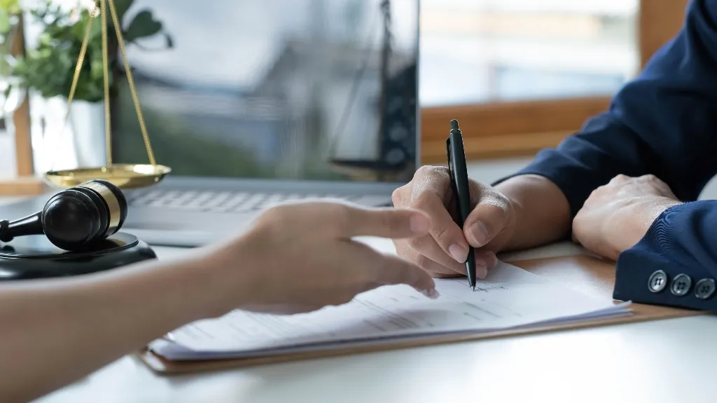 Close-up of two professionals reviewing and signing legal documents at a desk with a gavel and scales of justice.